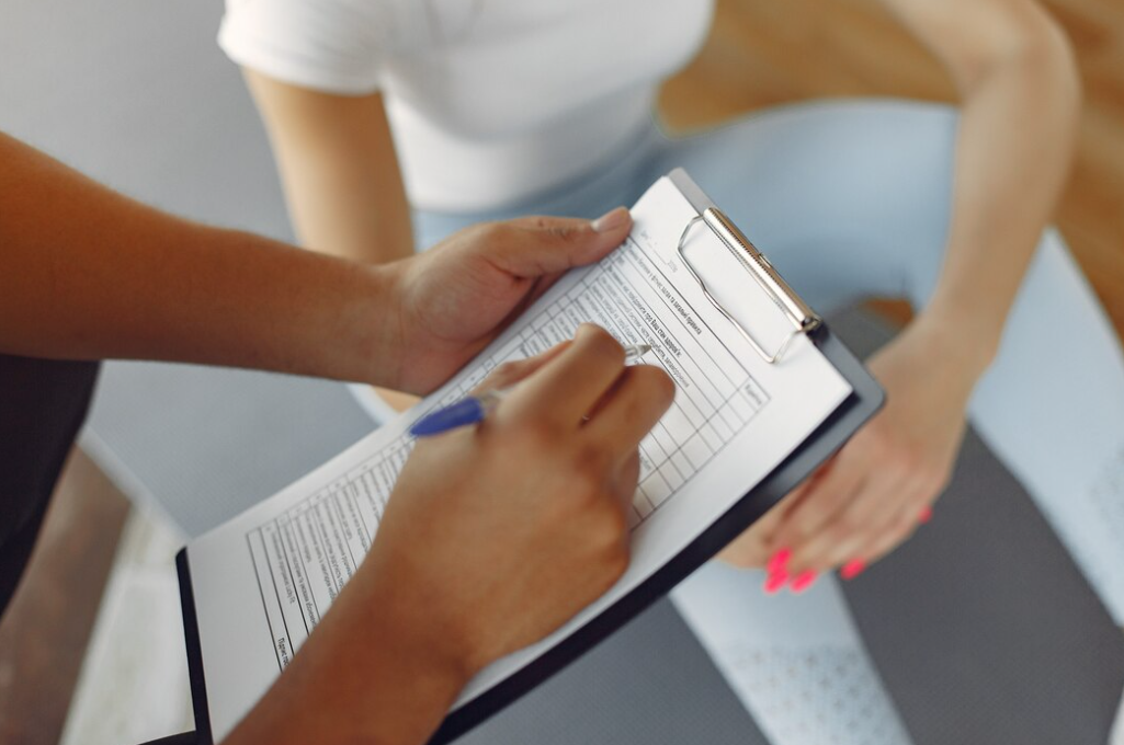 woman in blue leggings sitting on the chair, another person holds a sheet with info and makes notes