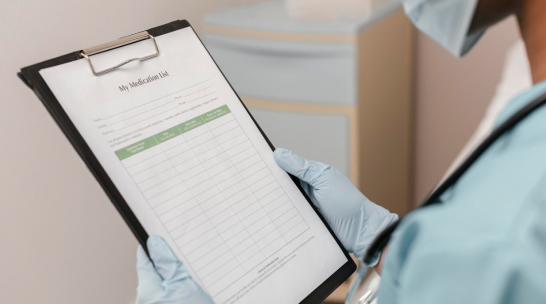 A person in a blue medical suit, mask, and gloves holds a sheet with my medication list on it