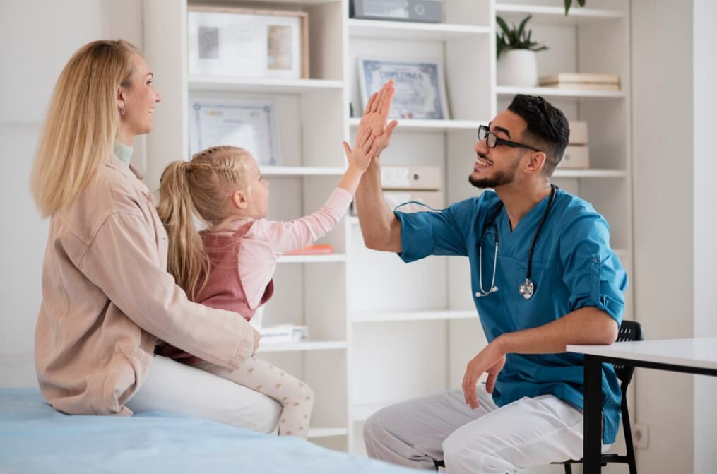 A young doctor high-fives a child with her mother watching in a clinic.