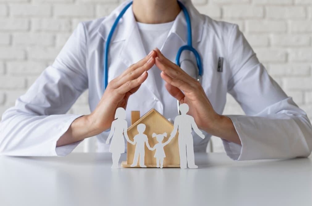 A doctor shields a paper family under a house, symbolizing healthcare protection.