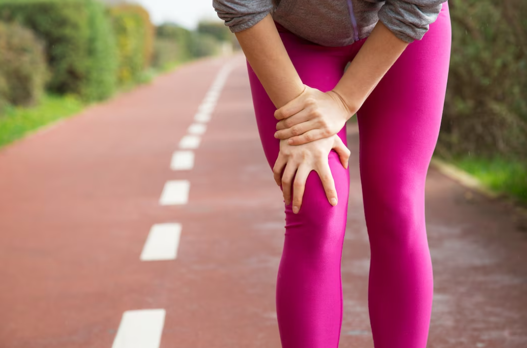 woman in pink leggings and a gray sweater holds her thigh on the running track