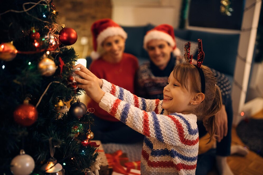 Image of a family decorating a Christmas tree, with the parents wearing Santa hats and the child wearing deer antlers.